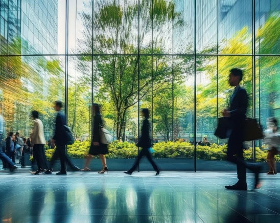 People walking inside modern office building with glass walls and trees outside.