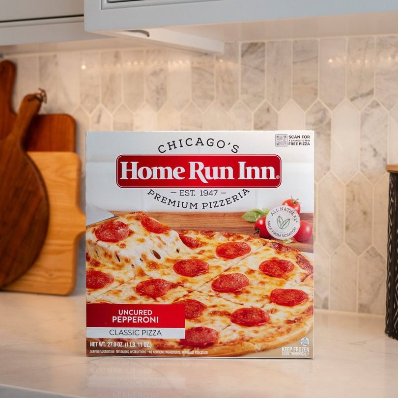 Pepperoni pizza box on kitchen countertop with chopping boards and decorative container.